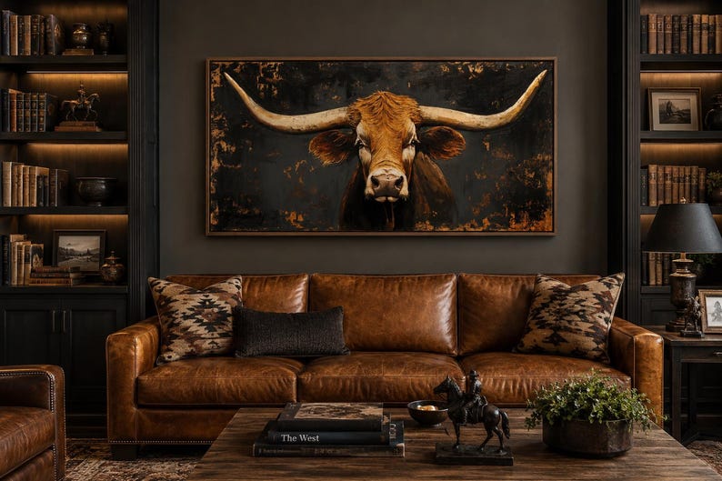 May include: A rectangular wall art piece featuring a longhorn cow portrait with large curved horns against a dark, textured background. The artwork is framed in a thin brown border and is centered above a brown leather sofa with patterned pillows.