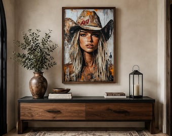 May include: A rectangular framed wall art print featuring a portrait of a person with long blonde hair wearing a wide-brimmed cowboy hat. The artwork uses a mix of neutral tones, orange accents, and paint splatter effects on a light background.