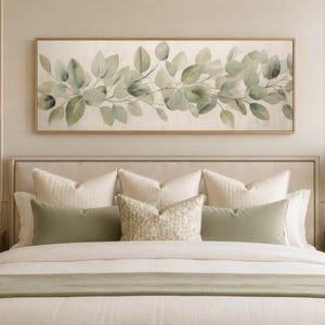 May include: A bedroom scene featuring a bed with cream-colored bedding, accented with sage green pillows and a matching bed runner. Above the bed hangs a rectangular artwork with a botanical print of green leaves. Two lamps flank the bed.