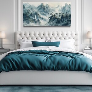 a serene and modern bedroom with a large bed, white tufted headboard, and a painting of a mountainous landscape above it. The bed is adorned with a blue comforter and white pillows, creating a tranquil and inviting atmosphere. The room is decorated with white walls, a gray rug, and two nightstands with lamps, adding to the overall elegance and sophistication of the space.