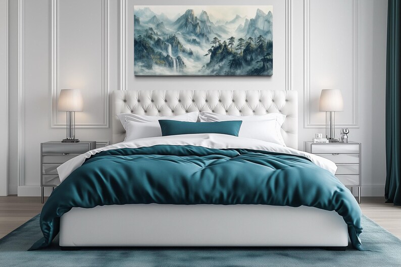 a serene and modern bedroom with a large bed, white tufted headboard, and a painting of a mountainous landscape above it. The bed is adorned with a blue comforter and white pillows, creating a tranquil and inviting atmosphere. The room is decorated with white walls, a gray rug, and two nightstands with lamps, adding to the overall elegance and sophistication of the space.