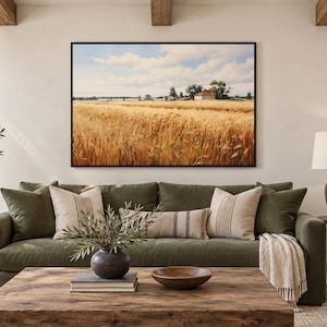 May include: A rectangular wall art print featuring a golden wheat field under a blue sky with white clouds, with a small house visible in the distance. The artwork is framed in a thin black border and displayed above a dark olive green sofa.
