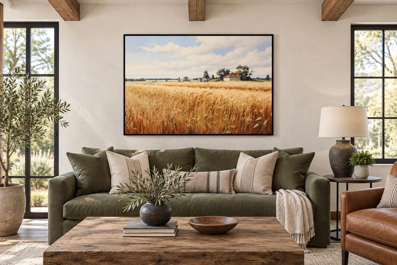 May include: A rectangular wall art print featuring a golden wheat field under a blue sky with white clouds, with a small house visible in the distance. The artwork is framed in a thin black border and displayed above a dark olive green sofa.
