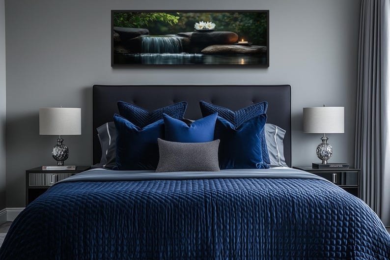 a serene bedroom scene with a large bed adorned with blue and gray bedding, flanked by two nightstands with lamps. Above the bed, a framed picture of a waterfall and flowers adds a touch of nature to the room's decor.
