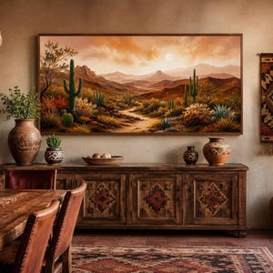 Op de afbeelding: Een rechthoekig landschapsschilderij met een woestijnscène met saguaro-cactussen, een kronkelend zandpad en verre bergen onder een warme oranje zonsonderganghemel, ingelijst in een dunne houten lijst boven een donkerhouten dressoir.