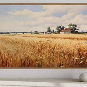 a serene rural landscape with a vast golden wheat field stretching out under a blue sky dotted with fluffy white clouds. In the distance, a small white house can be seen nestled among the trees. The scene is framed by a white border, and a white vase and a small white sculpture are placed on a white surface to the right of the frame.