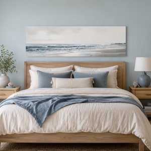 May include: A bedroom scene with a wooden bed frame, beige bedding, and blue accent pillows and throw. A seascape painting hangs above the bed. Two wooden nightstands flank the bed, each with a lamp. A neutral color palette creates a relaxing atmosphere.
