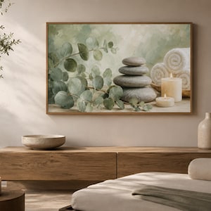 May include: A framed wall art print featuring a stack of smooth grey stones, green eucalyptus branches, white rolled towels, and two lit cream-coloured candles against a soft, muted green and beige painted background with a textured finish.