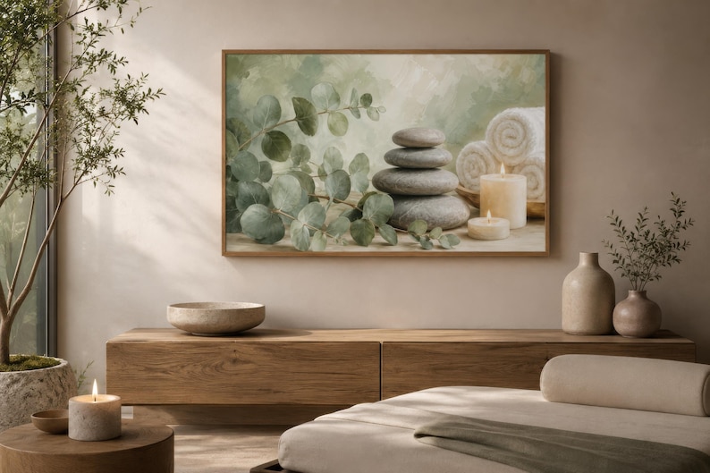 May include: A framed wall art print featuring a stack of smooth grey stones, green eucalyptus branches, white rolled towels, and two lit cream-coloured candles against a soft, muted green and beige painted background with a textured finish.
