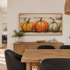 May include: A rectangular wall art piece featuring a painting of three pumpkins in orange, yellow, and muted green tones, set against a horizontal wood-plank background. The artwork is enclosed in a light-toned wooden frame and mounted on a wall.