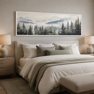 Puede incluir: Una pintura de paisaje panorámico en un marco blanco que presenta una cordillera brumosa y un denso bosque de árboles de hoja perenne en tonos azules, verdes y grises, exhibida horizontalmente sobre una cama tapizada de tonos neutros.