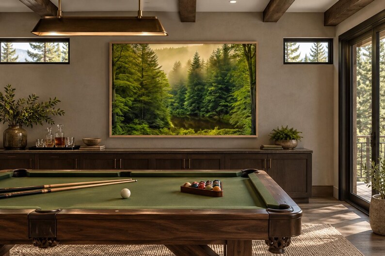 May include: A rectangular framed wall art print depicting a dense, misty green forest with tall pine trees and a dark pond. The artwork is mounted on a wall above a dark wooden cabinet and a green felt pool table with cues and balls.