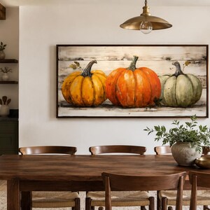 May include: A rectangular wall art piece featuring a painting of three pumpkins in yellow, orange, and muted green, set against a horizontal white wood plank background. The artwork is enclosed in a dark brown frame and displayed on a white wall.