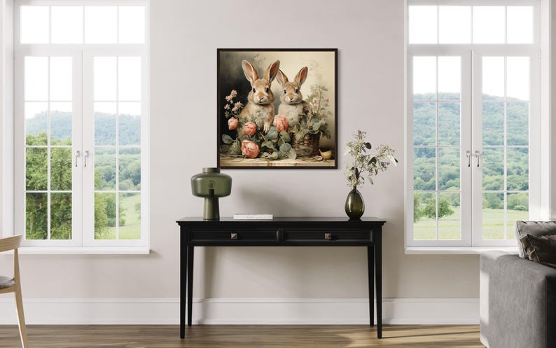 a living room with a black console table in the center, which has a vase, a lamp, and a book on it. Above the table, there is a framed painting of two rabbits sitting next to each other, surrounded by flowers. The room has large windows that provide a view of a green field and trees outside.