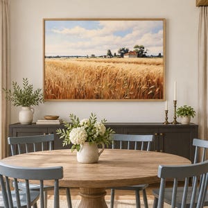 May include: A rectangular landscape painting in a light wood frame depicting a golden wheat field under a blue sky with white clouds, featuring a small house in the distance, displayed above a dark gray sideboard in a dining room setting.