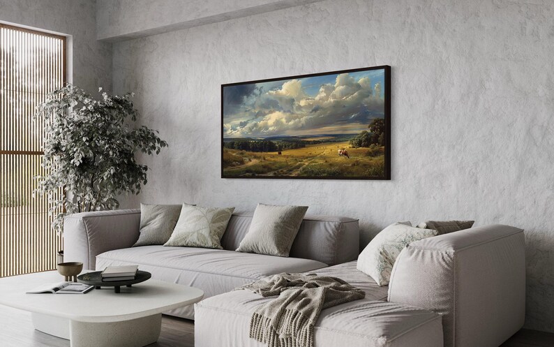 a living room with a large landscape painting hanging on the wall above a gray sofa. The painting features a countryside scene with a cloudy sky and rolling hills. The room has a minimalist and modern design, with a white coffee table and a potted plant adding a touch of greenery to the space.