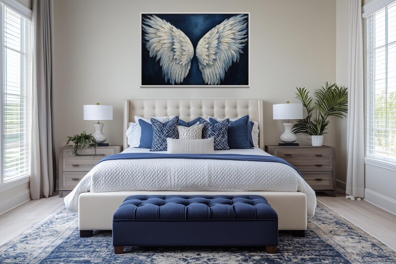 a well-decorated bedroom with a large bed, a tufted headboard, and a blue tufted ottoman. Above the bed is a large painting of white wings. The room also features a nightstand on each side of the bed, with a lamp and a plant. The walls are painted in a light color, and the floor is covered with a blue and white patterned rug.