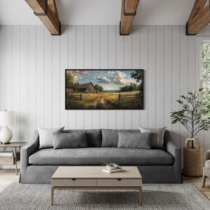 a cozy living room with a gray sofa, a wooden coffee table, and a large window overlooking a rural landscape. A framed painting of a farm scene hangs on the wall, adding a touch of nature to the space.