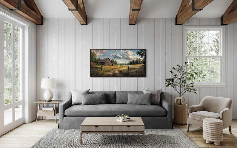 a cozy living room with a gray sofa, a wooden coffee table, and a large window overlooking a rural landscape. A framed painting of a farm scene hangs on the wall, adding a touch of nature to the space.