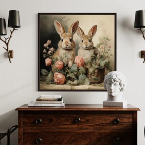 a wooden dresser with a framed painting of two rabbits sitting in a basket of flowers hanging on the wall above it. The dresser also has a statue of a man's head and some books on top.