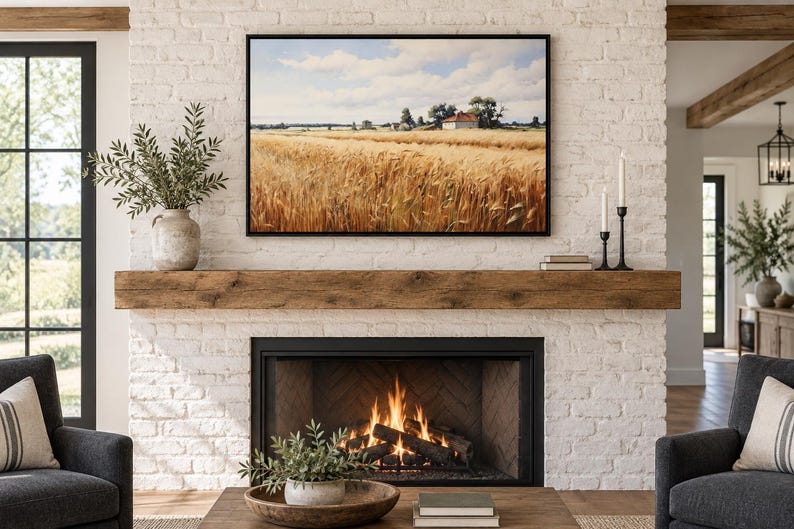 May include: A rectangular landscape wall art print featuring a golden wheat field under a blue sky with white clouds, displayed in a thin black frame above a rustic wooden fireplace mantel and a white brick fireplace with a burning fire inside.