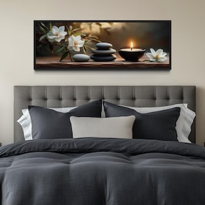 a serene bedroom scene with a gray tufted headboard, a gray comforter, and a gray throw pillow. Above the bed, there is a framed artwork featuring a still life of stones, flowers, and a lit candle. The room has a neutral color palette with a touch of greenery from the potted plant and vases.