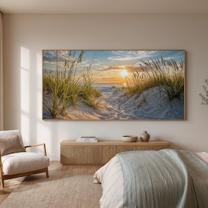 May include: A large rectangular wall art print featuring a coastal sunset scene with sand dunes, tall beach grass, and a bright sun low on the horizon. The artwork is framed in a light wood border and displayed above a wooden media console in a room.