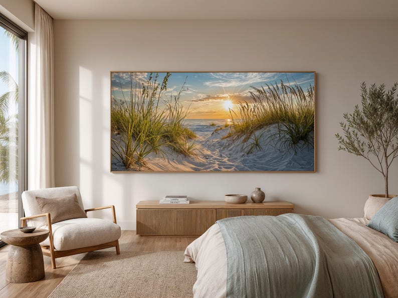 May include: A large rectangular wall art print featuring a coastal sunset scene with sand dunes, tall beach grass, and a bright sun low on the horizon. The artwork is framed in a light wood border and displayed above a wooden media console in a room.