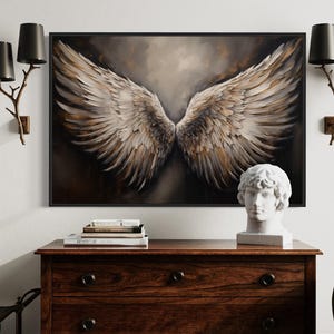 a large framed painting of a pair of angel wings hanging on a wall above a wooden dresser. The dresser has a bust of a person and some books on top of it. There are also two black lamps on either side of the painting.