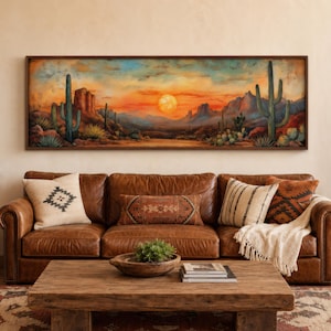 May include: A panoramic rectangular wall art piece featuring a desert landscape at sunset with a bright orange sun, saguaro cacti, and rocky mountains. The artwork is framed in dark wood and hangs above a brown leather sofa with decorative pillows.