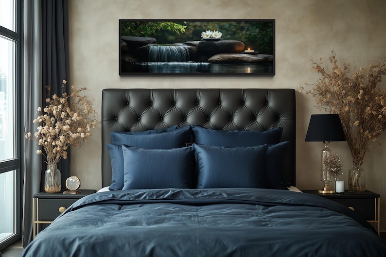 a serene bedroom scene with a large bed adorned with blue bedding and a tufted headboard. Above the bed, a framed picture of a waterfall and flowers adds a touch of nature to the room. The bed is positioned next to a nightstand with a lamp and a vase of flowers, creating a cozy and inviting atmosphere.