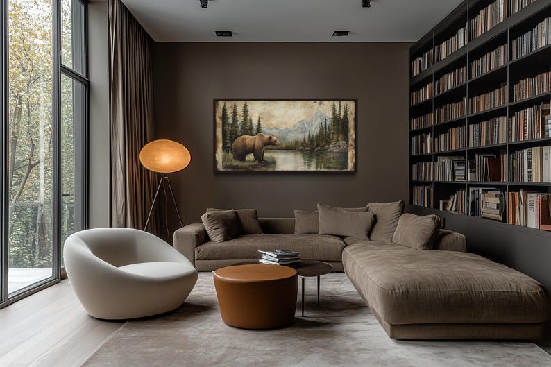 a cozy living room with a large window overlooking a scenic outdoor view. The room is furnished with a comfortable sofa, a round coffee table, and a floor lamp. A large bookshelf filled with books is also present, creating a warm and inviting atmosphere. A framed painting of a bear in a forest setting adds a touch of nature and artistry to the space.