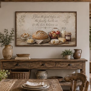 Op de afbeelding: Een rustieke eetkamer met een houten tafel en een ingelijst kunstwerk. Het kunstwerk toont een stilleven van brood, kaas, fruit en zuivelproducten. De tekst op het kunstwerk luidt: "Zegen het voedsel voor ons, de familie naast ons, & de liefde tussen ons."