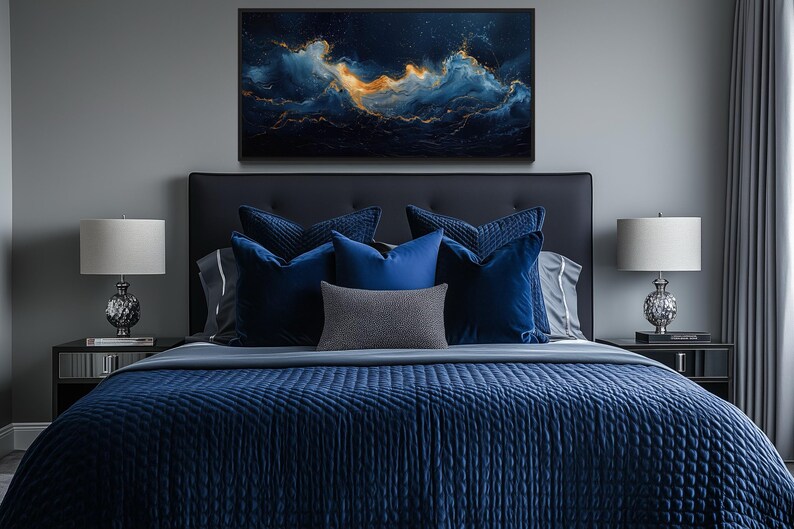a luxurious bedroom with a large bed adorned with blue and gray bedding, flanked by two nightstands with lamps. Above the bed, a large framed artwork of a night sky with swirling clouds and stars is displayed on the wall.