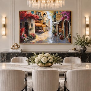May include: A rectangular oil painting in a gold frame depicting a sunlit European street scene with vibrant red and pink flowers, outdoor cafe seating, and stone architecture, displayed on a wall above a dark marble console table and dining set.