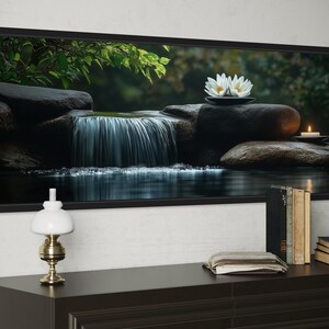 A framed photograph of a serene waterfall and lotus flowers is displayed on a wall above a wooden dresser with a lamp, books, and a clock.