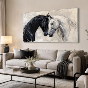 Puede incluir: Obra de arte en lienzo enmarcada que representa dos caballos, uno negro y otro blanco, con sus cabezas juntas. La obra cuelga sobre un sofá beige en un salón, creando un punto focal.