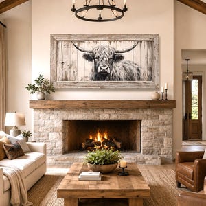 May include: A rectangular wall art piece featuring a black and white illustration of a longhorn cow on a vertical wood plank background, enclosed in a weathered light gray wooden frame, mounted above a stone fireplace in a living room.