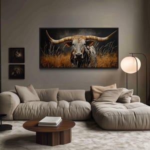 a modern living room with a large, comfortable-looking couch and a coffee table. Above the couch, there is a large framed painting of a bull with long horns. The room also features a bookshelf filled with books and a floor lamp.