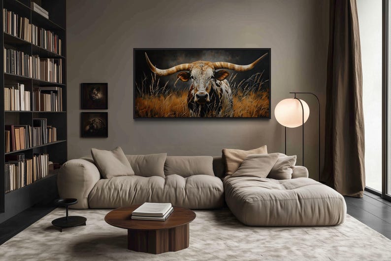 a modern living room with a large, comfortable-looking couch and a coffee table. Above the couch, there is a large framed painting of a bull with long horns. The room also features a bookshelf filled with books and a floor lamp.