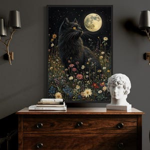 a black cat sitting in a field of flowers, with a full moon in the background. The cat is surrounded by various flowers and plants, creating a serene and peaceful scene. The image is framed and displayed on a wooden dresser, which also has a statue and some books on it. The dresser is situated in a room with a gray wall and two wall sconces.