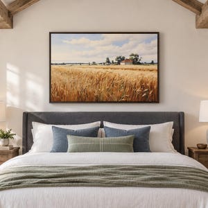 May include: A rectangular landscape wall art print featuring a golden wheat field under a blue sky with white clouds, displayed in a dark brown frame above a dark gray upholstered bed with white bedding, blue pillows, and a green throw blanket.