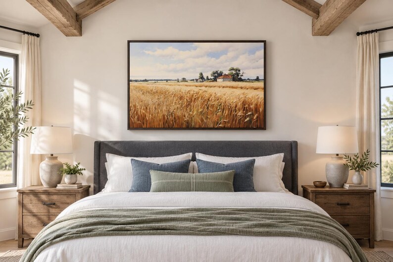 May include: A rectangular landscape wall art print featuring a golden wheat field under a blue sky with white clouds, displayed in a dark brown frame above a dark gray upholstered bed with white bedding, blue pillows, and a green throw blanket.