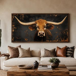 May include: A rectangular wall art piece featuring a detailed painting of a brown longhorn steer with large, sweeping horns against a dark, textured background. The artwork is enclosed in a simple dark wood frame and hangs above a beige sofa.