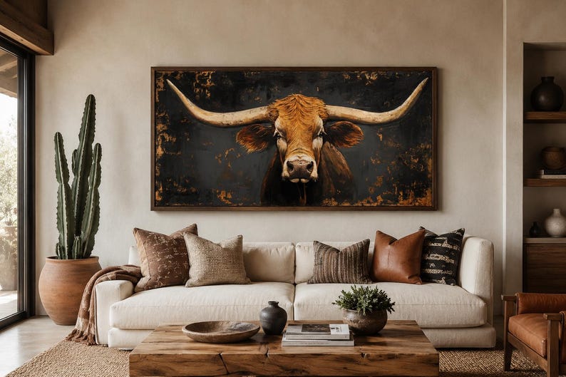 May include: A rectangular wall art piece featuring a detailed painting of a brown longhorn steer with large, sweeping horns against a dark, textured background. The artwork is enclosed in a simple dark wood frame and hangs above a beige sofa.