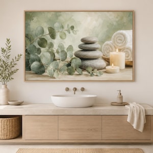 May include: A rectangular wall art print in a light wood frame featuring a painted scene of a stack of four smooth grey stones, green eucalyptus branches, two white pillar candles, and three rolled white towels against a soft green background.