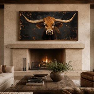 May include: A rectangular wall art piece featuring a longhorn cow with large, curved horns centered against a dark, textured background with hints of gold, displayed in a wooden frame above a stone fireplace in a living room setting.