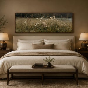 May include: A bedroom featuring a beige upholstered bed with a landscape painting of a field of white flowers above the headboard. The room includes two dark wood nightstands with lamps, a wooden bench at the foot of the bed, and a tall potted plant.