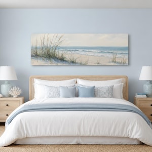 Könnte beinhalten: Ein rechteckiges Landschaftsgemälde einer Küstenszene mit Sanddünen, hohem Strandgras und blauen Meereswellen, das über einem Bett mit weißer Bettwäsche, einer hellblauen Tagesdecke und zwei passenden hellblauen Nachttischlampen hängt.