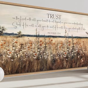 Op de afbeelding: Een rechthoekig ingelijst wandkunstwerk met een landschap van een goudkleurig veld met witte en oranje wilde bloemen onder een zachte lucht. De tekst luidt: TRUST in the Lord with all your heart; do not depend on your own understanding. Seek His will in all you do, and He will show you which path to take. PROVERBS 3:5-6.